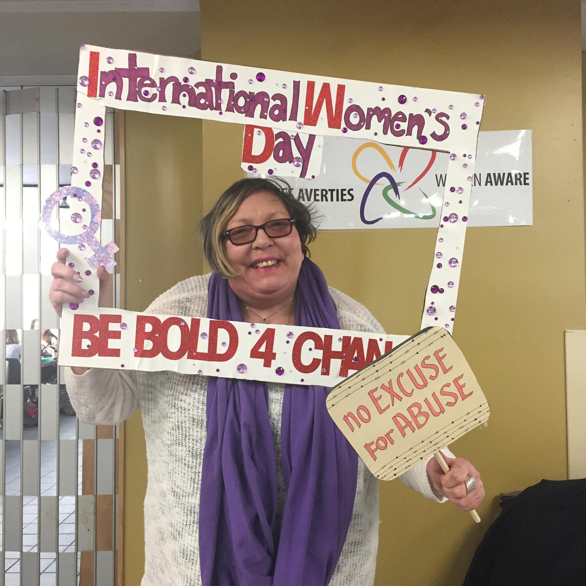 June is standing and holding up a sign abour Internaitonal Women's Day that says "Be Bold 4 Change"